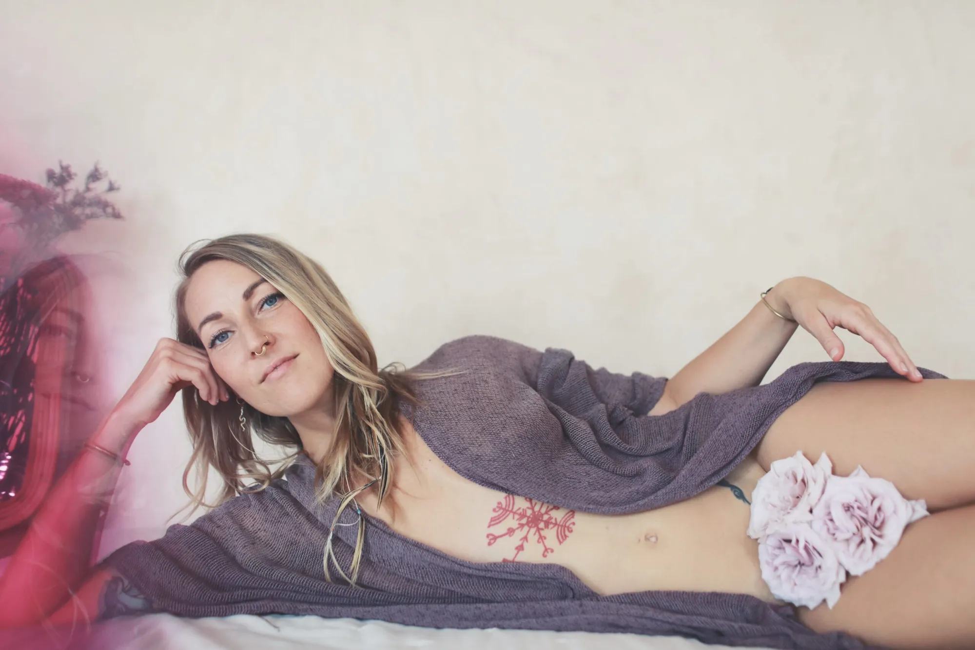 Linda Kaarina lying on her side wrapped in a soft knitted throw, roses resting beside her, looking into the camera